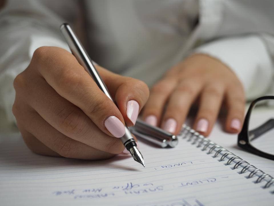 A hand writing a client's name and a lunch time into a lined notebook, beside a pair of glasses.